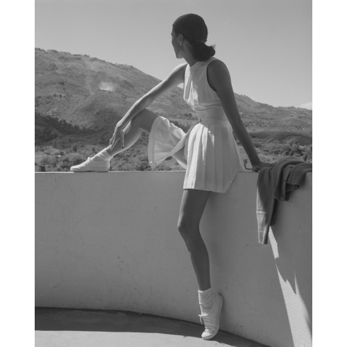 Woman Wearing Tennis Outfit, Seated On Wall, With One Leg On Top Of Wall, Looking At The...