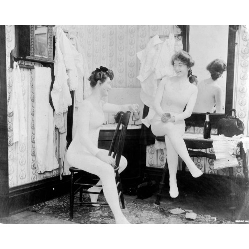 Two Young Women, Wearing Long Underclothing, One Seated On Chair And Smoking, The Other Seated...